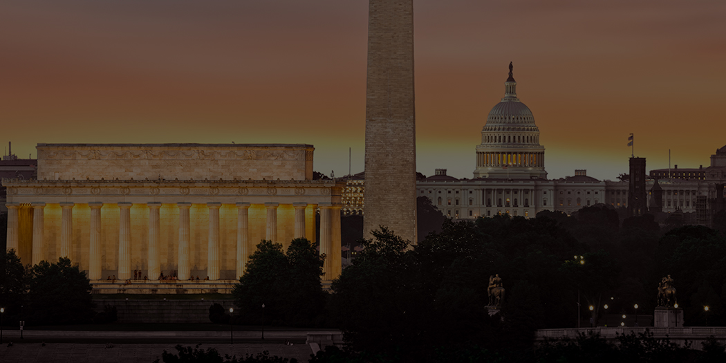 A photo of Washington DC at dawn; the Lincoln Memorial is in the foreground, with the Washington Monument and U.S. Capitol behind it. The sun is rising in the distance, just behind the capital.