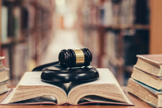 A photo of a gavel sitting on an open law book, the stacks of a library out of focus in the background.