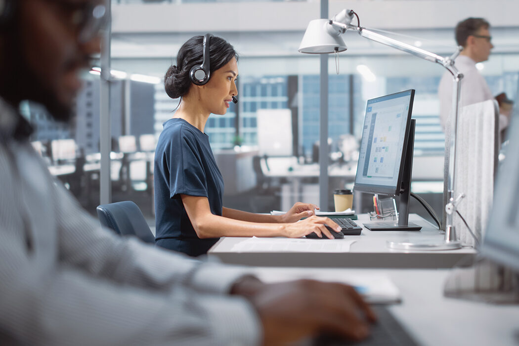 A photo of an Asian woman wearing a headset and working on a computer in an open-air office. In the bottom left corner in the foreground, there is also an out-of-focus African American man.