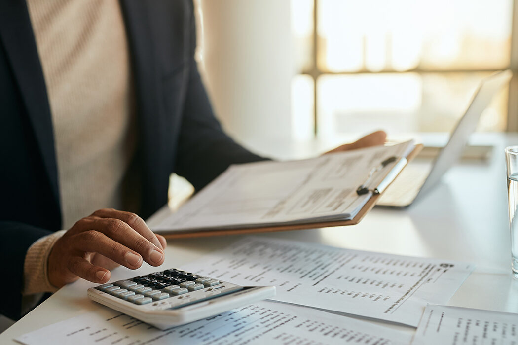 A black woman uses a calculator and examines a report while sitting at a desk covered in financial papers. The crop only includes her torso and arms.