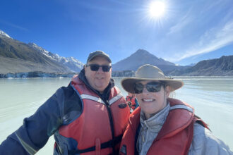 A photo of NOSSCR CEO Steve Gardner and his wife Cheryl Levine in a boat on a lake nestled between mountains. There is a bright blue sky, and the sun is visible; Steve and Cheryl are both wearing life jackets, hats, and sunglasses.