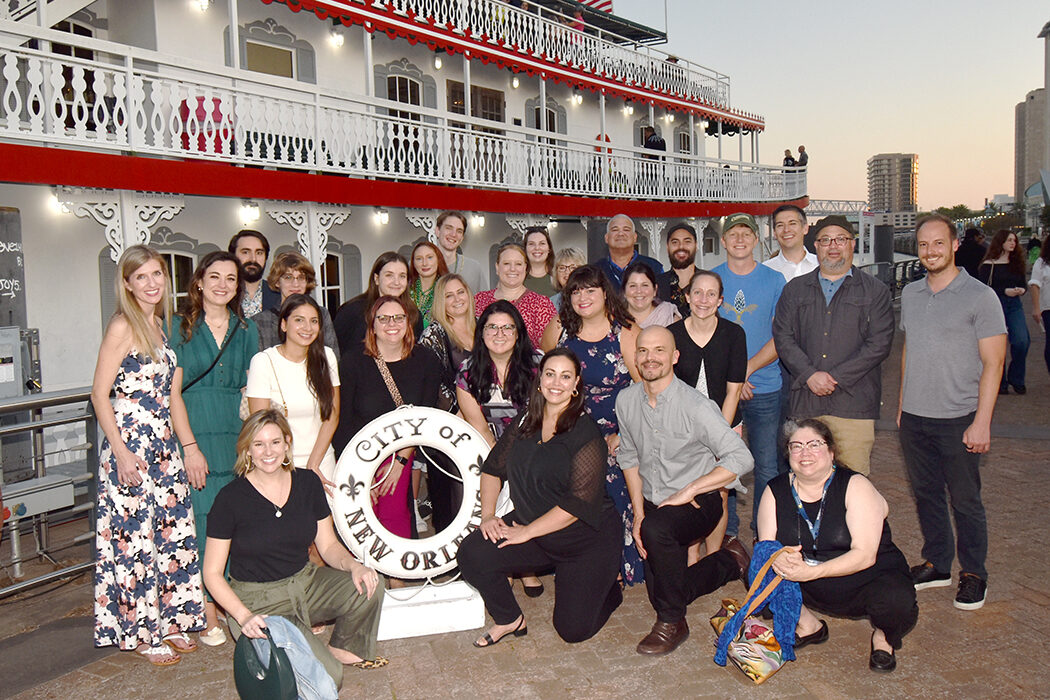 2023 NextGen attendees are posing on a dock in front of a red and white riverboat. Attendees are standing and kneeling to fit everyone in, and they are surrounding a statue in the shape of a life preserver wit the words "City of New Orleans" emblazoned on it.