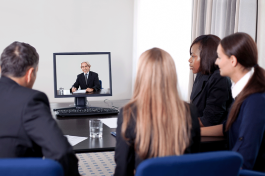 Four young people in suits sit in front of a video screen displaying an older man in a suit.